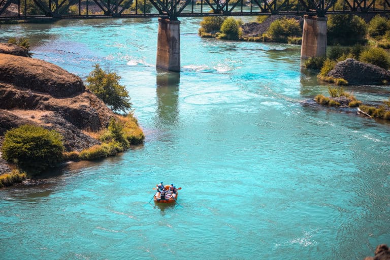 A red rafting boat with a group of people floating down a calm river with turquoise blue water. There is a bridge behind them with green shrubs and rocks surrounding the river.