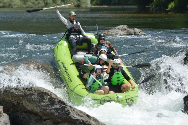 A group of six people in helmets and life jackets laughing and cheering on a bright green raft navigating whitewater rapids