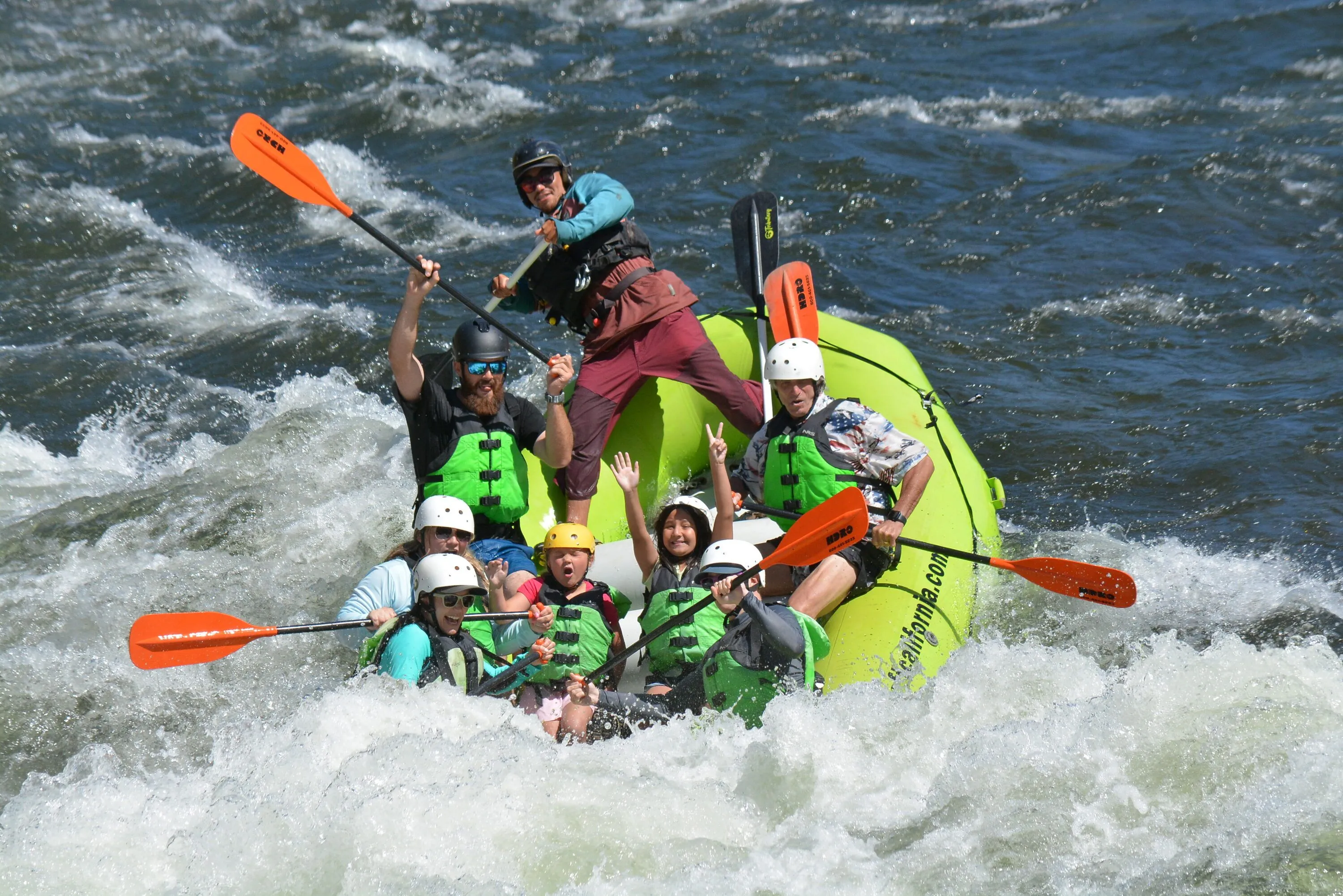 Eight cheering rafters with orange paddles raised and a guide standing mid-raft on a green Raft California raft crashing through whitewater