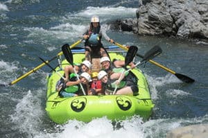 A group of people including kids in a rafting boat paddling through whitewater rapids with rocks surrounding the river.