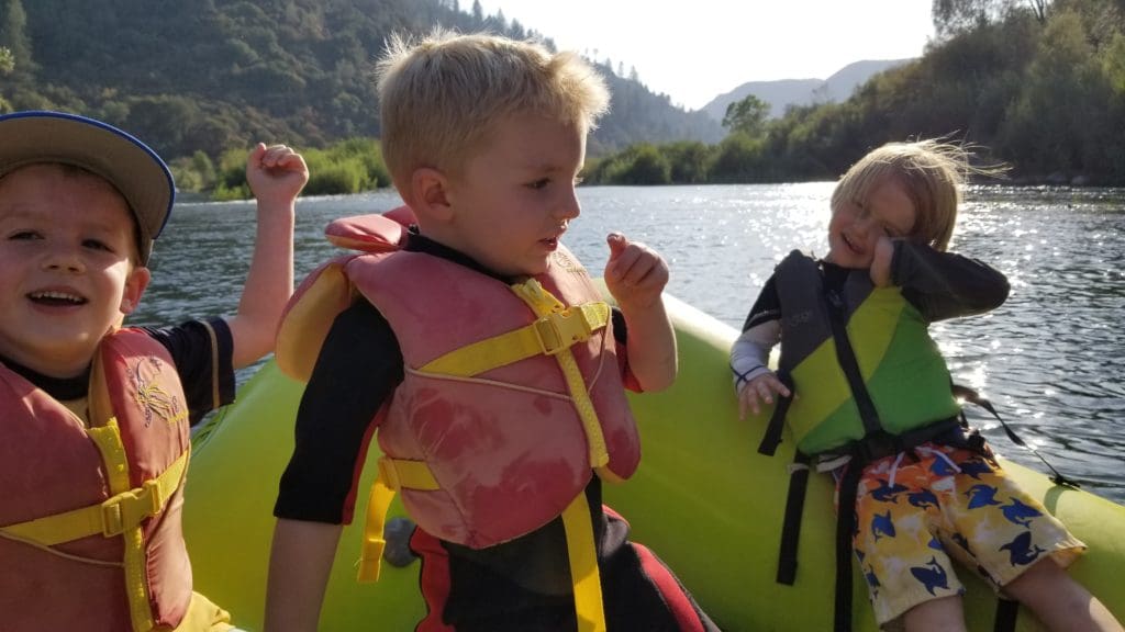 Three young children sitting on a rafting boat with a calm river and green trees in the background.