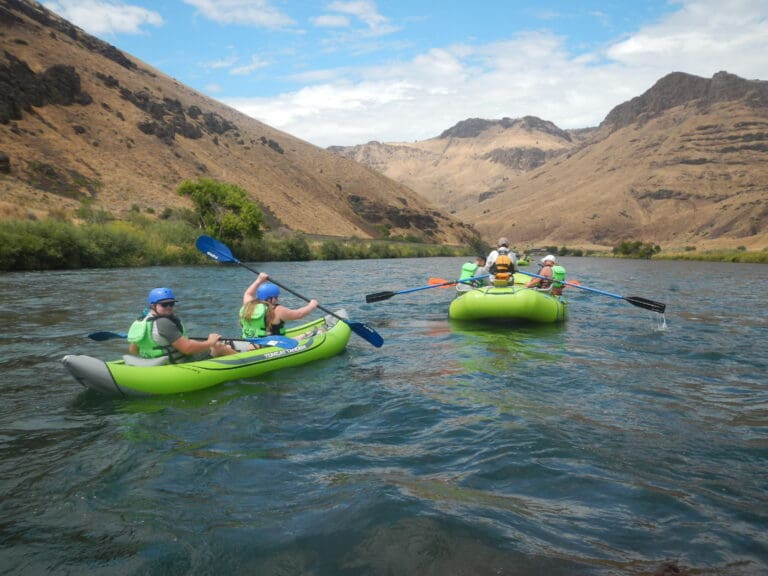 People in a rafting boat and a kayak paddle down a calm river in a canyon with brown grassy hillsides.