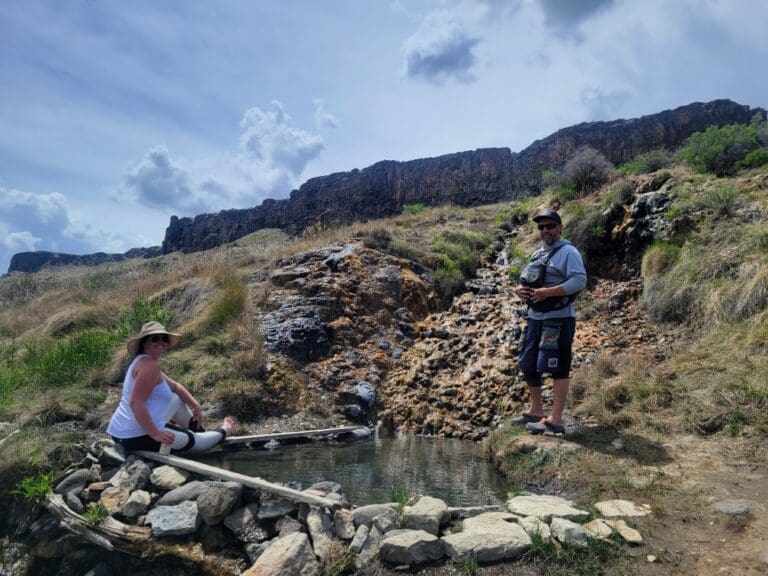 Two people sitting and standing next to a small hot spring with a grassy hillside and large rock wall in the background.