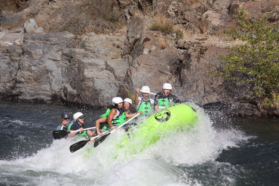 A group of people in a rafting boat in the middle of a whitewater rapid. The bow of the boat is sticking up in the air. Large rock walls in the background.