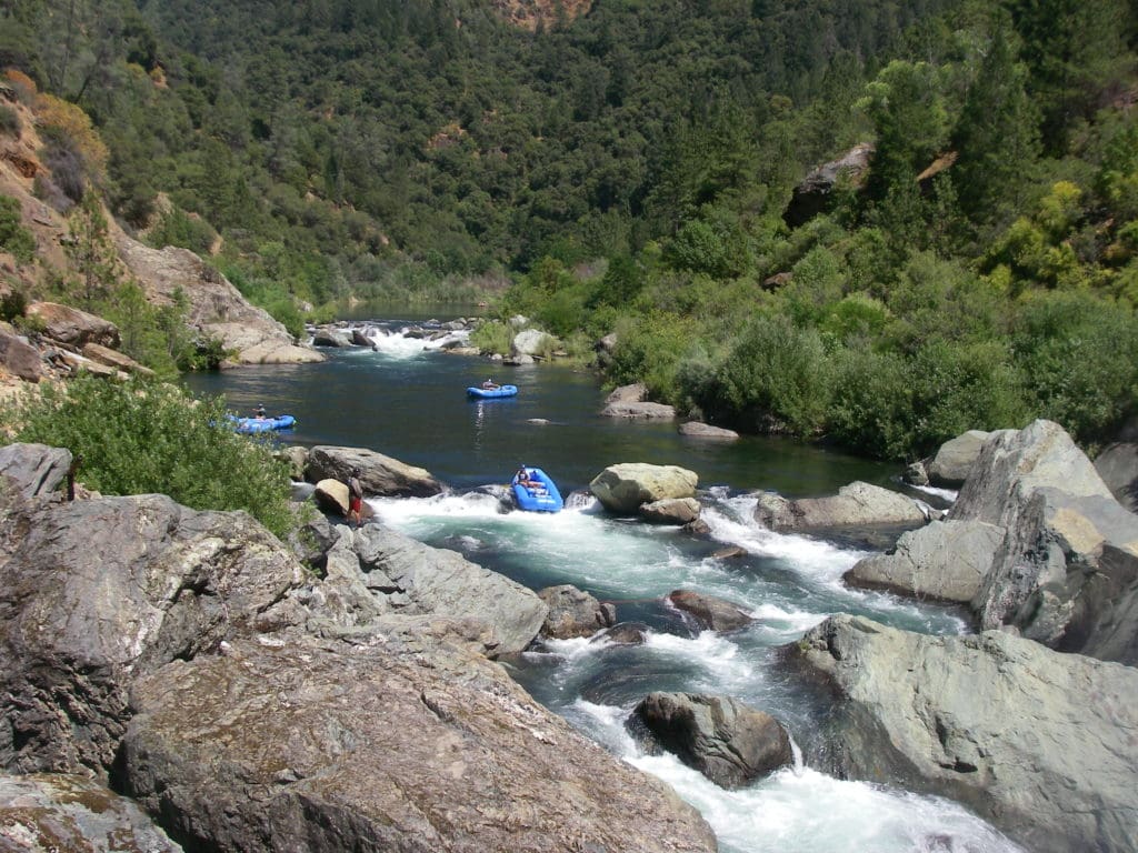 Three rafting boats paddling through a river with large boulders and whitewater rapids. Green forest and another whitewater rapid in the background