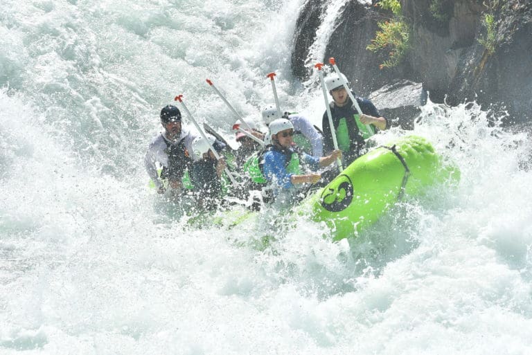 A group of people in a green rafting boat in the middle of a large whitewater rapid. Large rocks in the background.