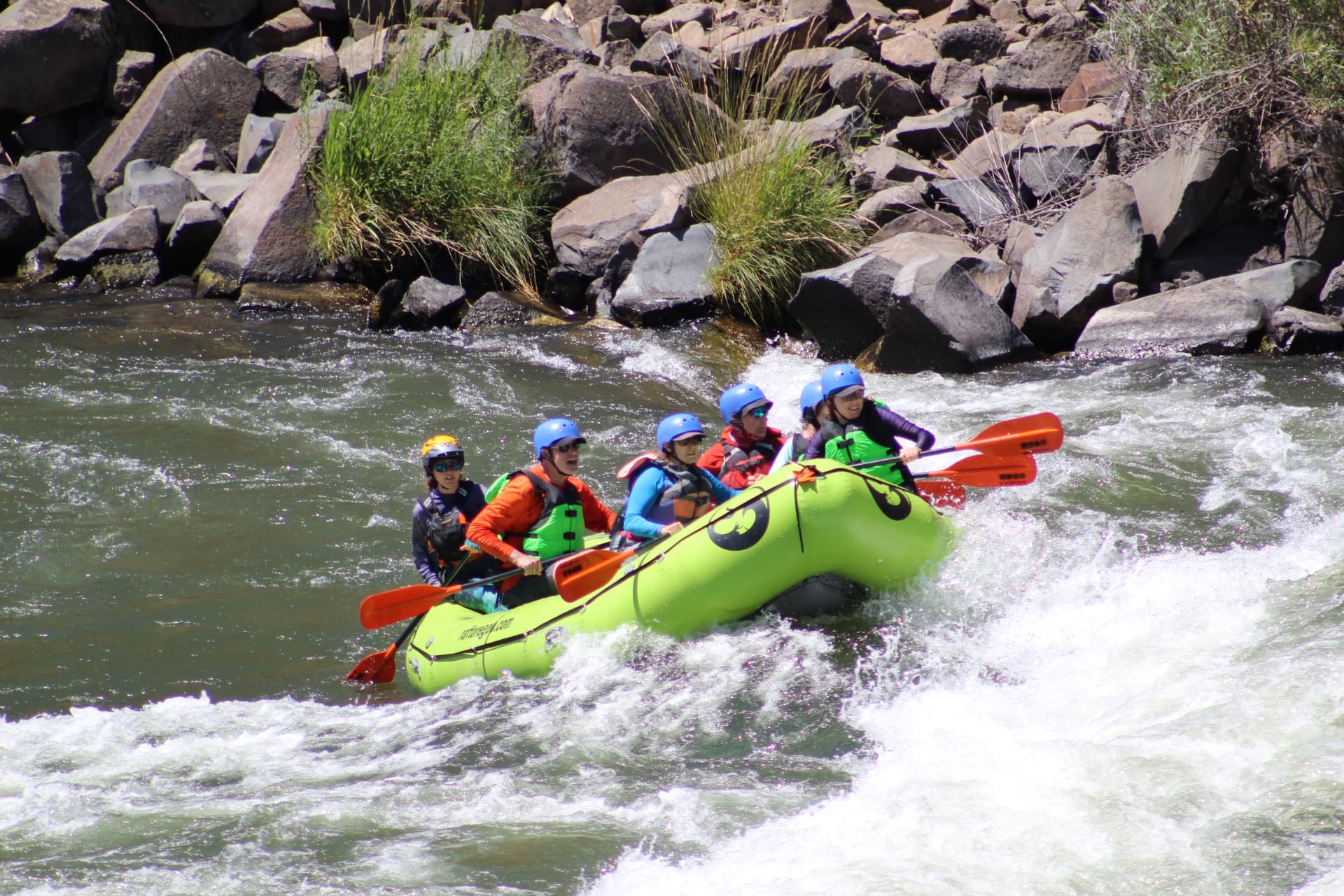 A group of people on a rafting boat paddling through a whitewater rapid on a river surrounded by rocks and green grass.