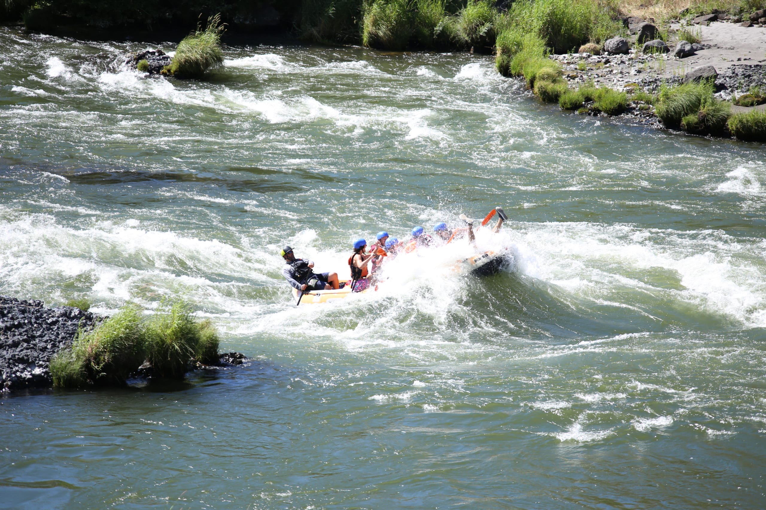 A group of people on a rafting boat almost fully submerged in a whitewater rapid on a river surrounded by rocks and green grass.