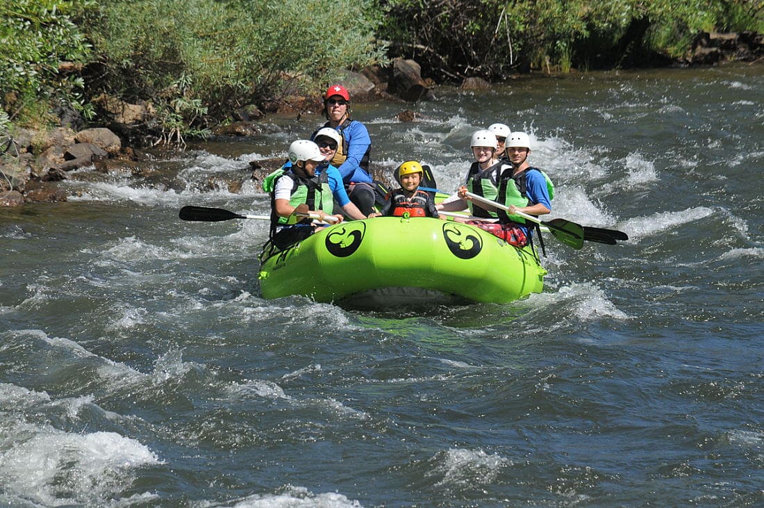 A family including kids are paddling in a rafting boat through small whitewater waves on a river surrounded by green shrubs.