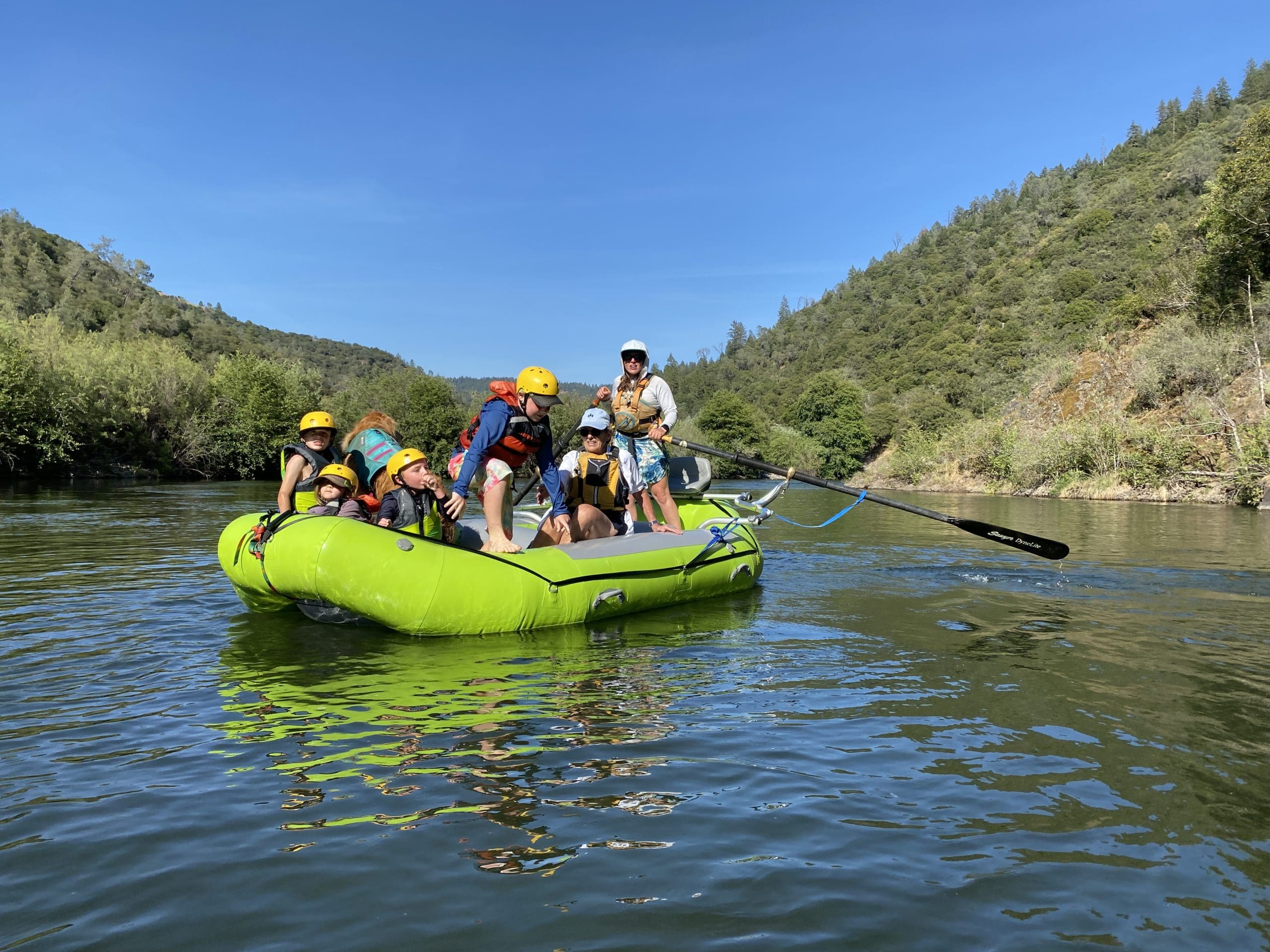 A group of people, including kids, are sitting on a rafting boat on a calm river. One kids looks like he's about to jump into the water front the boat.