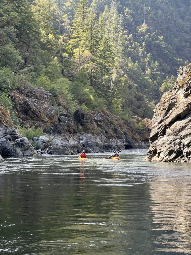 Two kayakers paddle down a calm river with rock canyons above them.
