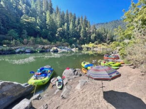 Whitewater rafting boats tied up on the banks of a calm river. There is an umbrella open on the beach.