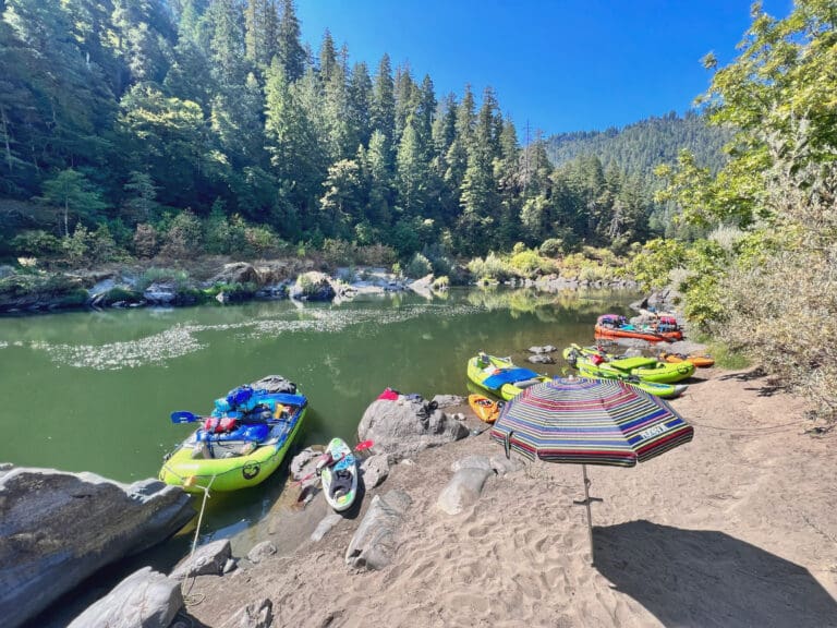 Whitewater rafting boats tied up on the banks of a calm river. There is an umbrella open on the beach.