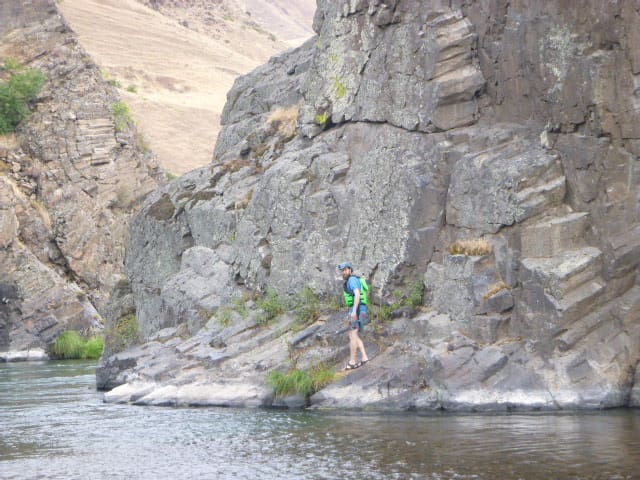 A person standing in the distance on a rock next to a calm river in a rocky canyon.