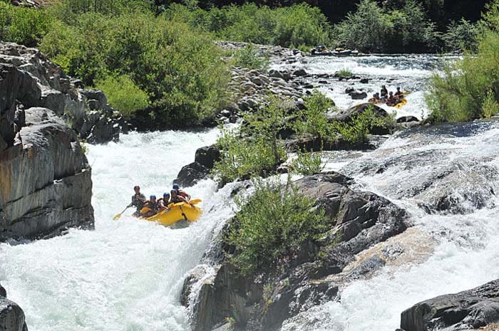 A rafting boat full of people navigate a large whitewater rapid with tall rocks surrounding the boat, making a narrow chute. Another rafting boat full of people are behind them on the river. Green bushes line the river.