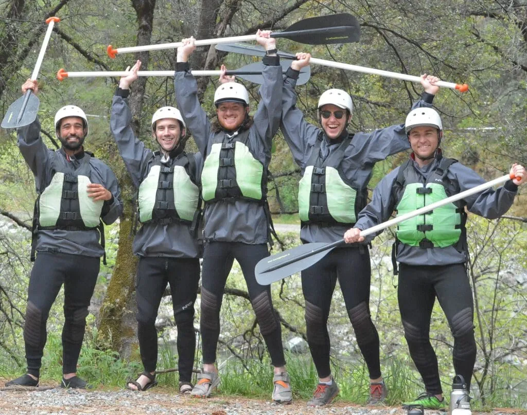 A group of paddlers wearing wetsuits, lifejackets and helmets, holding paddles next to a calm river.