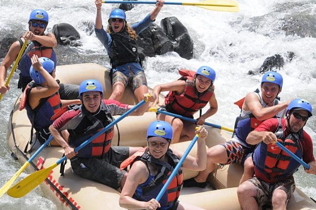 A group of people in a rafting boat smiling and holding their paddles up. A river with whitewater rapids and boulders in the background.