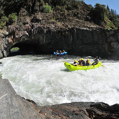 Two rafting boats full of people paddle through whitewater rapids. Rock walls and a cave are in the background.