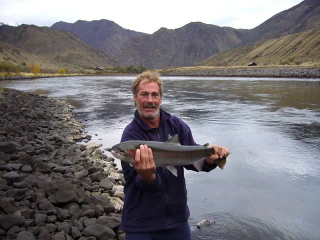 A man standing with a medium sized fish in his hands. A calm river, a rocky shore and brown mountains in the background.