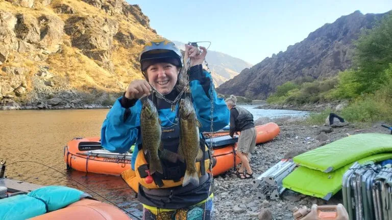 A person smiling holding up two small fishes. There are rafting boats tied up on the shore of a calm river. A rocky canyon in the background.