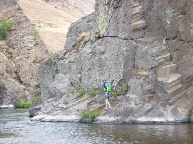 A person standing in the distance on a rock next to a calm river in a rocky canyon.