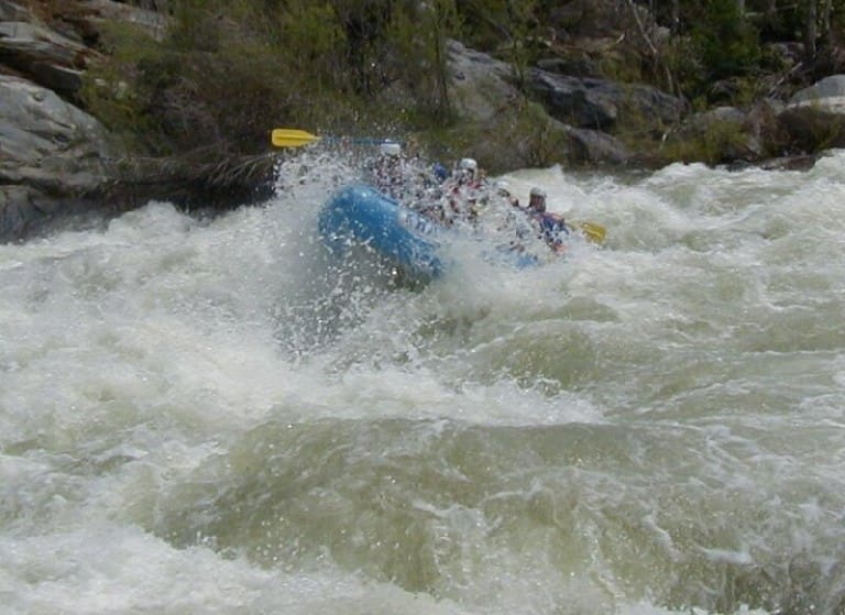 A rafting boat full of people navigate a large whitewater rapid. The boat is partially submerged and the river water is splashing the people. Large rocks and green trees in the background.