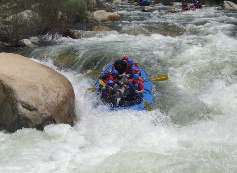 A group of people in a rafting boat navigate a large whitewater rapid. The bow of the boat is partially submerged in the river. There is a large boulder in the water next to the boat. More rocks and another rafting boat in the background.