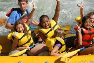Children in a rafting boat holding paddles, smiling with their arms in the air. A river with whitewater splashing behind them.