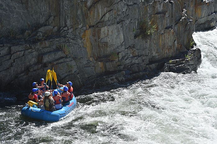A group of people in a rafting boat holding their paddles together in the air for a "paddle high five" large rock wall in the background.
