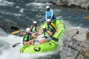 A group of people in a green rafting boat navigating a whitewater rapid. There are rocks surrounding the river.