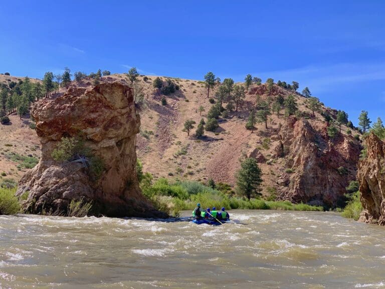 A rafting boat on a river with several people in it. Red rocks in the background.