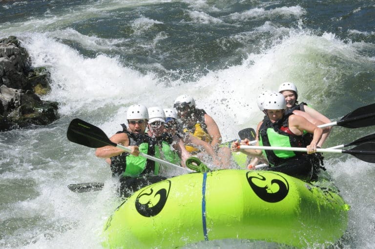 A group of people in a rafting boat paddling through a large whitewater rapid. The river water is splashing them. Rocks in the river in the background.