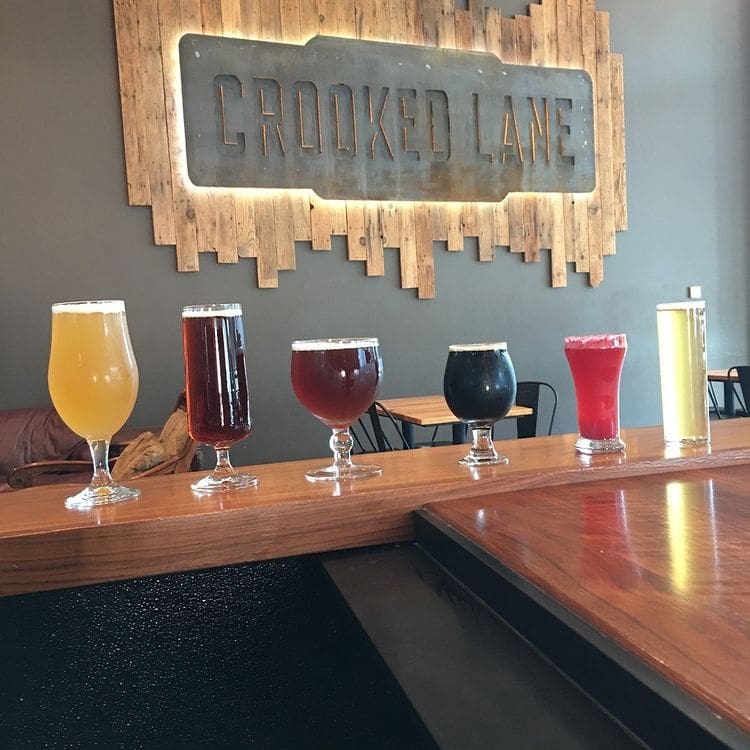 Six glasses of various sizes filled with beer of various colors. A sign in the background says "Crooked Lane"
