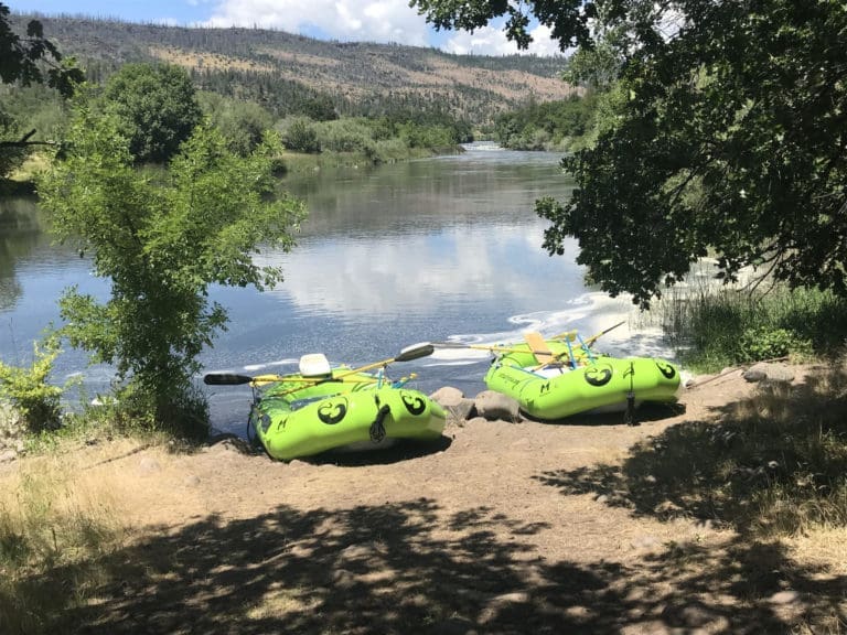 Two green inflatable rafting boats tied on the shore of a calm river with a forested hillside in the background.