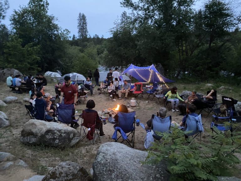 A group of people sitting around a campfire with tents and a green forest in the background.