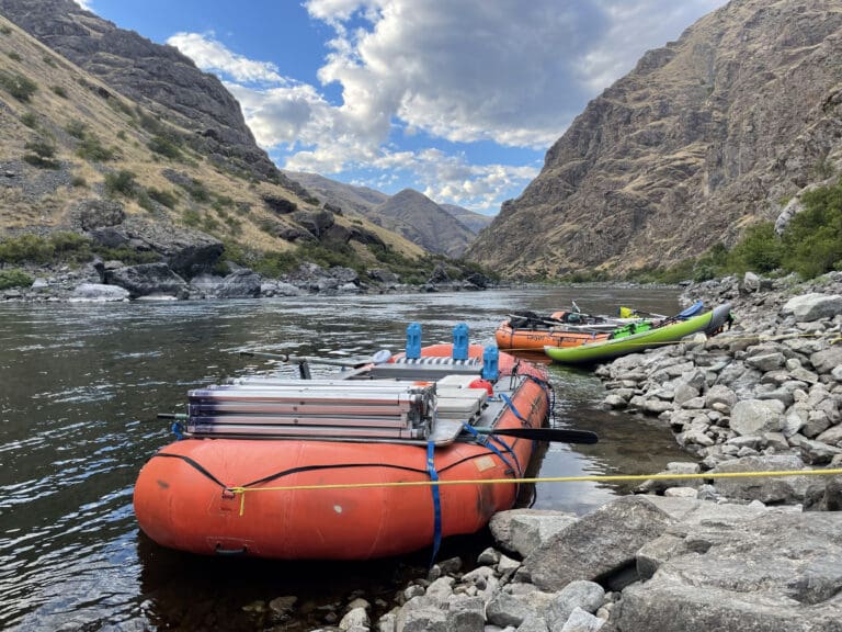 Orange white water rafting boat tied up to rocky river shore in a river canyon.
