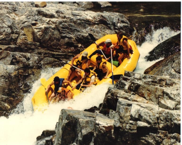 A yellow rafting boat full of people paddling down a steep waterfall surrounded by rocks.