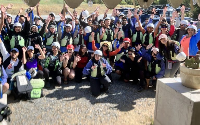 Large group of rafters with helmets and lifejackets on with their hands up in the air.
