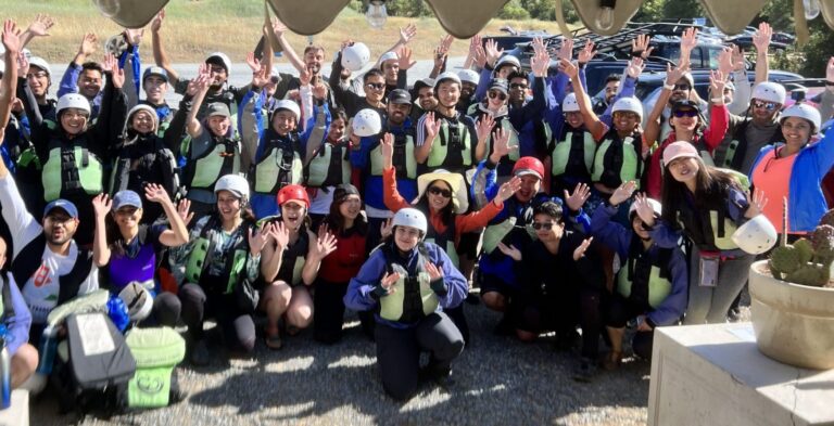 Large group of rafters with helmets and lifejackets on with their hands up in the air.