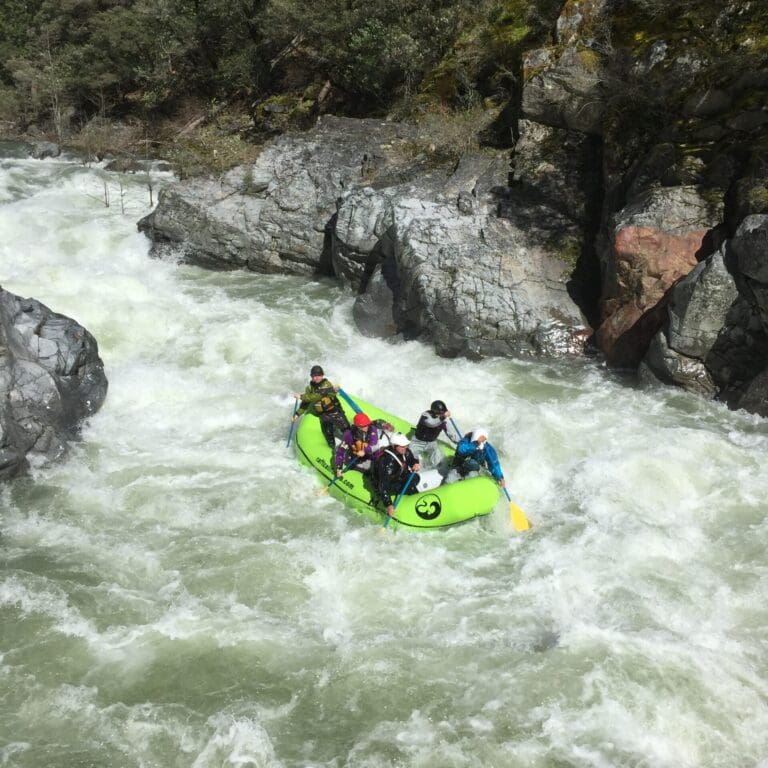 A rafting boat filled with people paddling through whitewater on a narrow river in a rocky canyon.