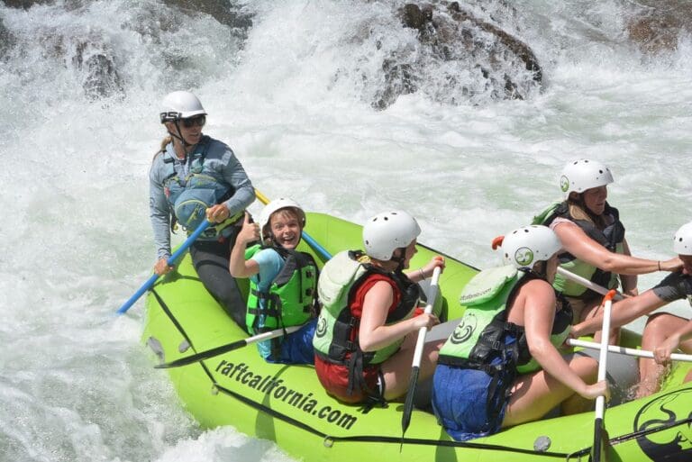 A group of people including kids in a rafting boat paddling through a large whitewater rapid. There are large rocks in the river behind them.