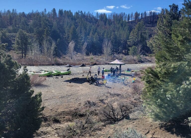 A campsite next to a river with green rafting boats tied on shore. There are people standing under a pop up tent and chairs set up around a fire pit.