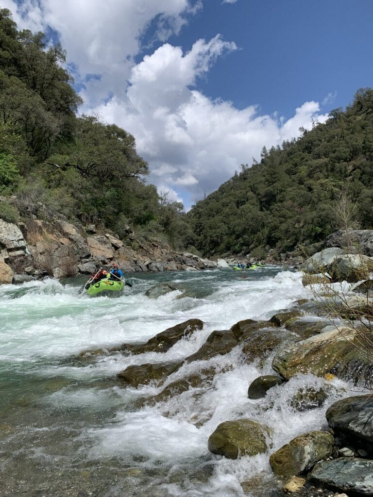 A group of people in a rafting boat paddling down whitewater rapids on a river in a lush green canyon. A few rafting boats full of people are behind them.