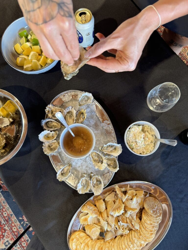 Two hands holding an oyster in it's shell over a table filled with platters of other food including a platter of oysters on ice, a bowl of lemons and limes, a platter of crackers and smoked fish, a beer can and an empty wine glass.