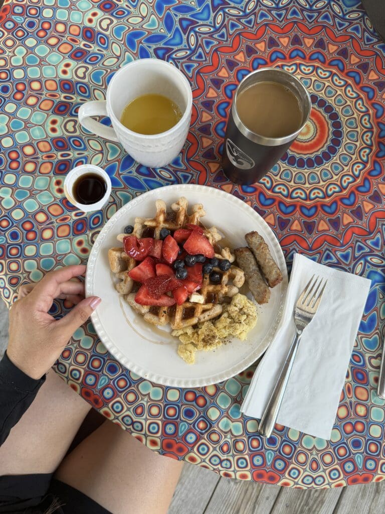 A table with a colorful tablecloth with a place of waffles topped with berries with a side of eggs and sausage. Three cups with beverages sit around the plate.