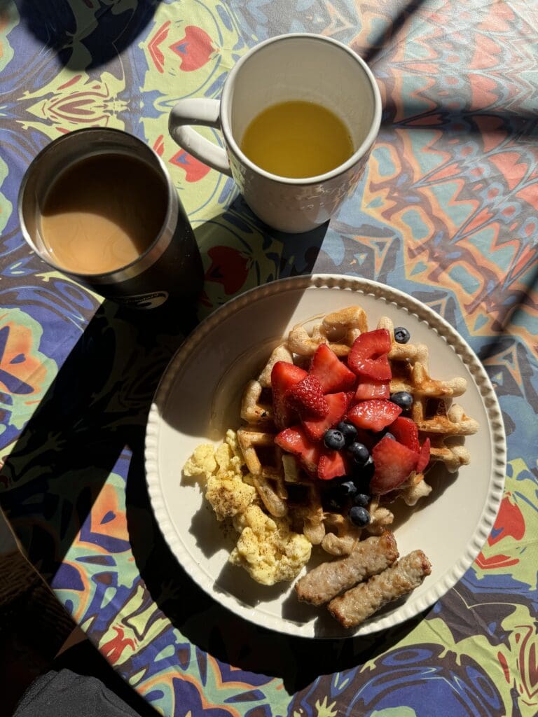 A table with a plate of waffles topped with berries with a side of eggs and sausage. Two cups with beverages in them sit on top a colorful tablecloth.