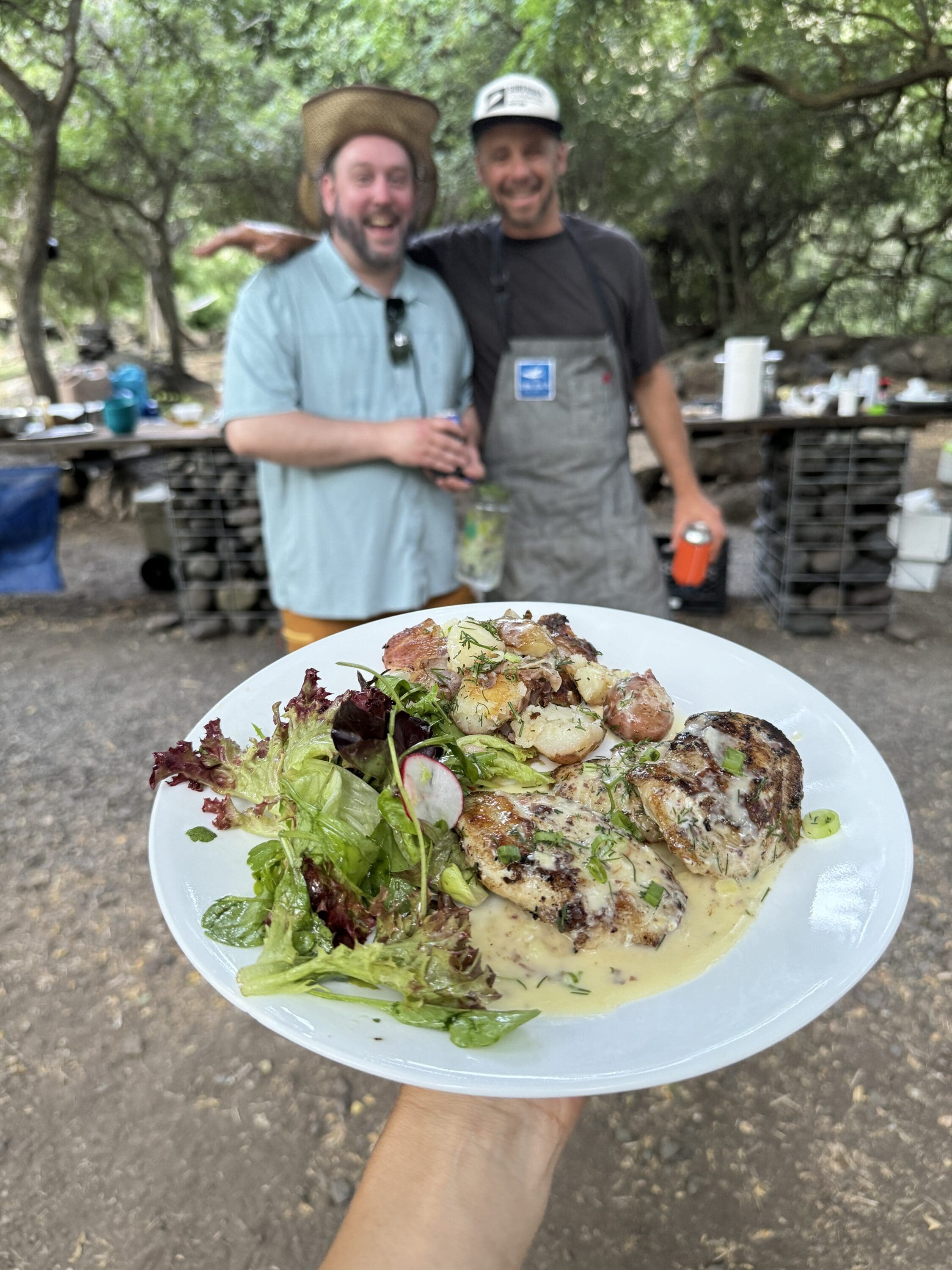 A plate of chicken, potatoes and salad. Two people in the background are holding drinks and have their arms around each other.