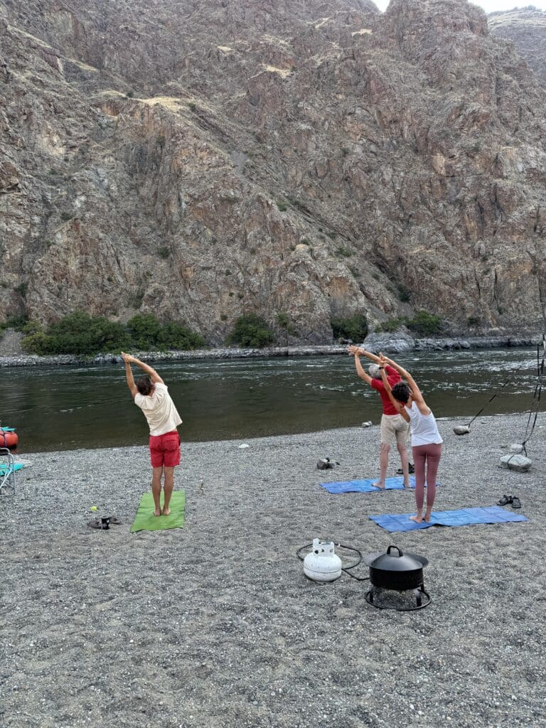 Three people doing yoga on the shore of the Snake River with a large rockface on the opposite side of the river. The people are standing on colorful mats while doing a yoga pose.