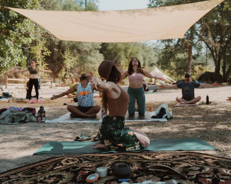 People on yoga mats doing various poses. The mats are sitting on dirt with green trees behind them.