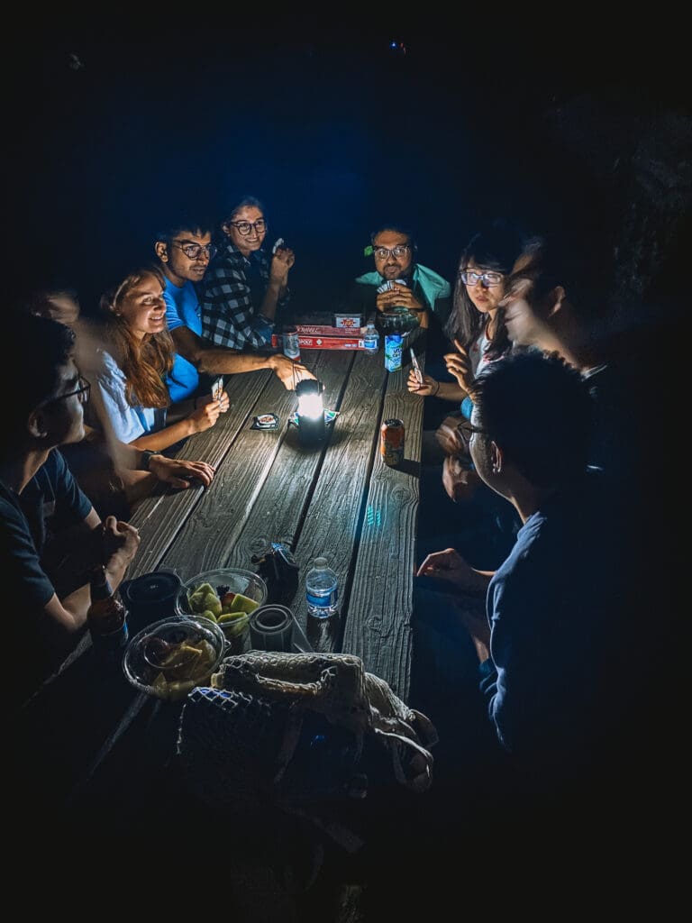 A group of people sitting around a wooden picnic table at night with a lantern, drinks and games.